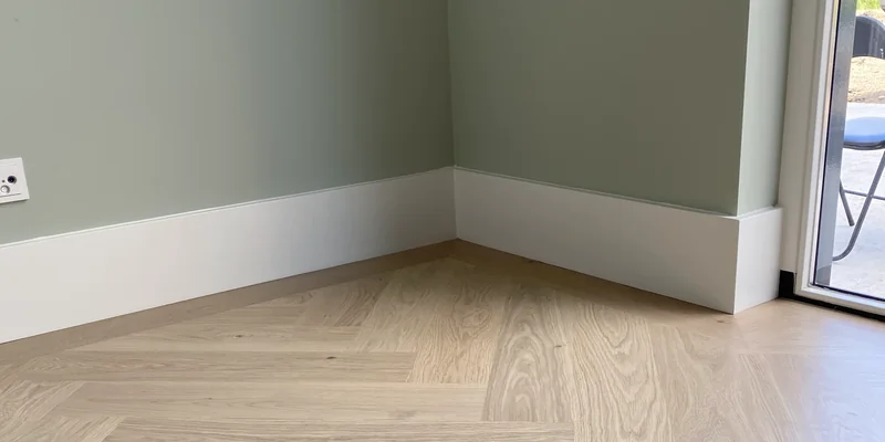 Witte gladde plinten in een hoek met groene muur en visgraatvloer, strak tegen de muur geplaatst
